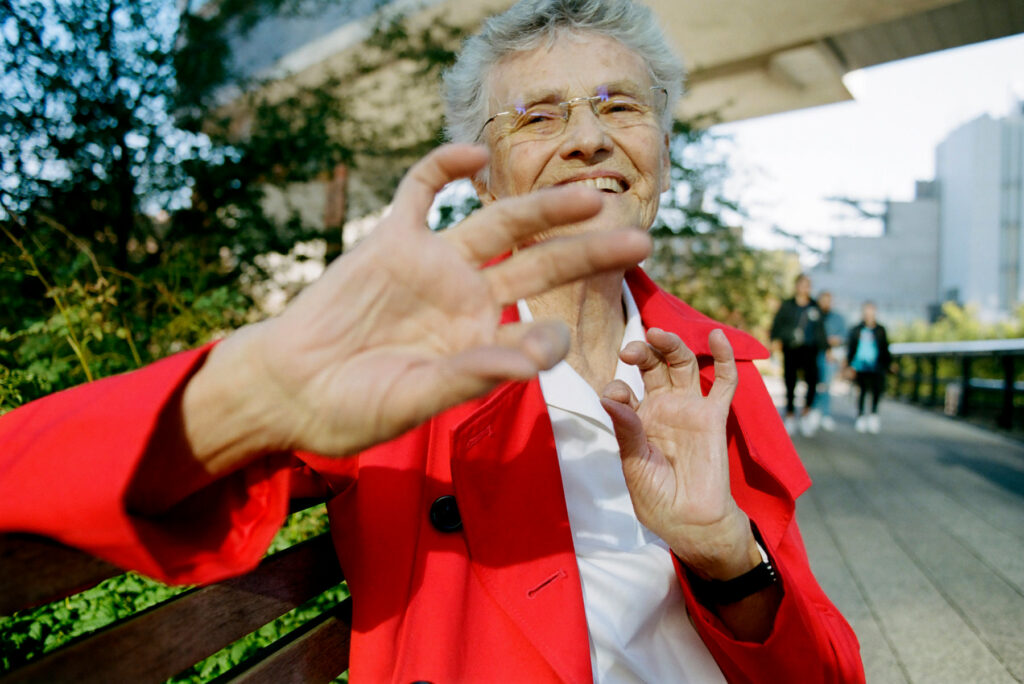 woman with grey hair and red jacket holding her hand up at the camera