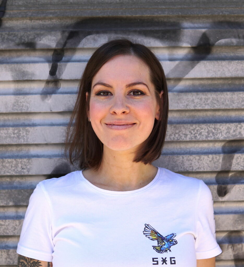 A young woman smiles facing the camera, with a white t shirt. She stands in front of a graffitied roller door.